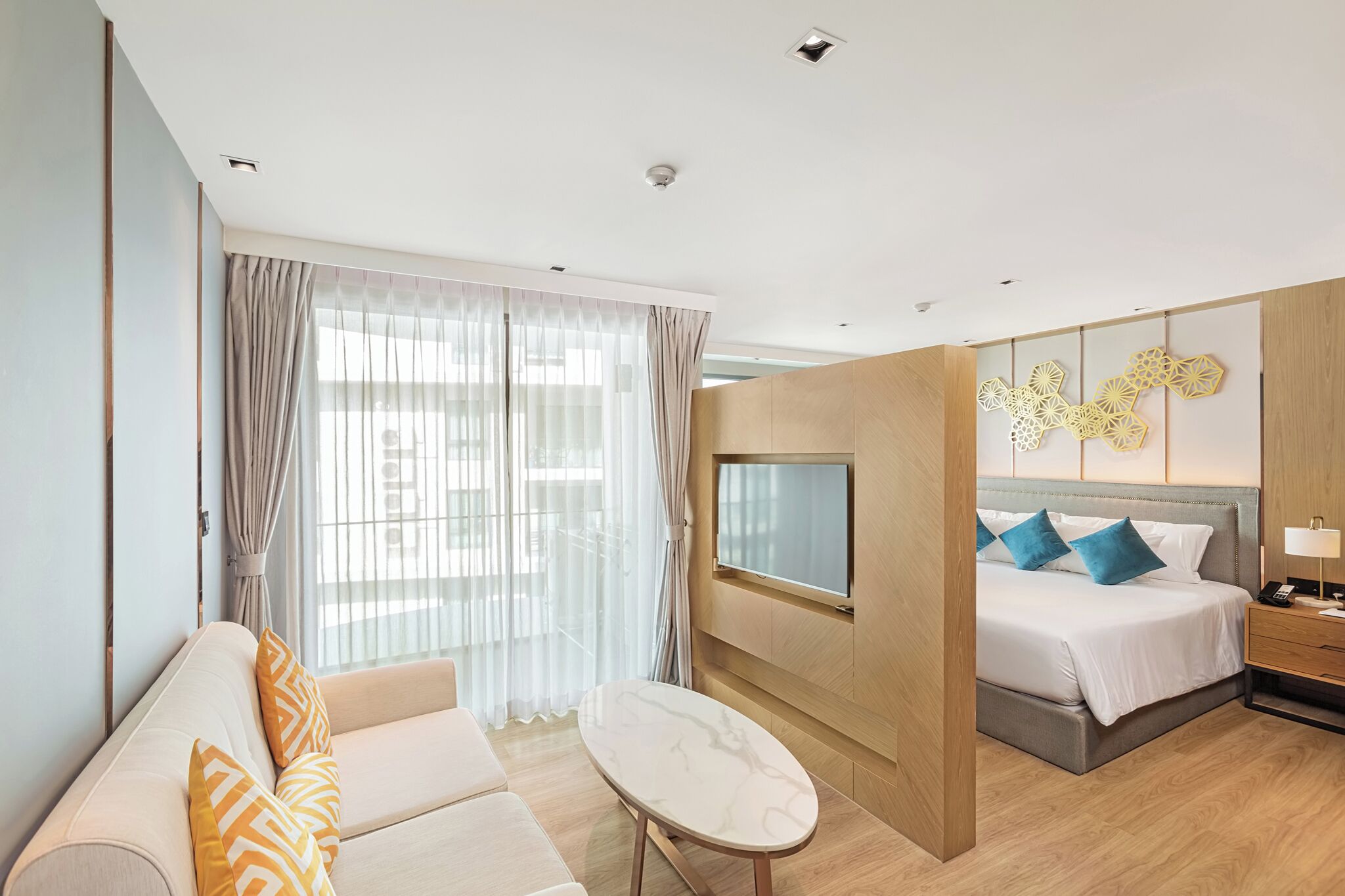 A modern hotel room with a sofa, a small table, a bed with blue pillows, a TV, and a decorative wall piece.