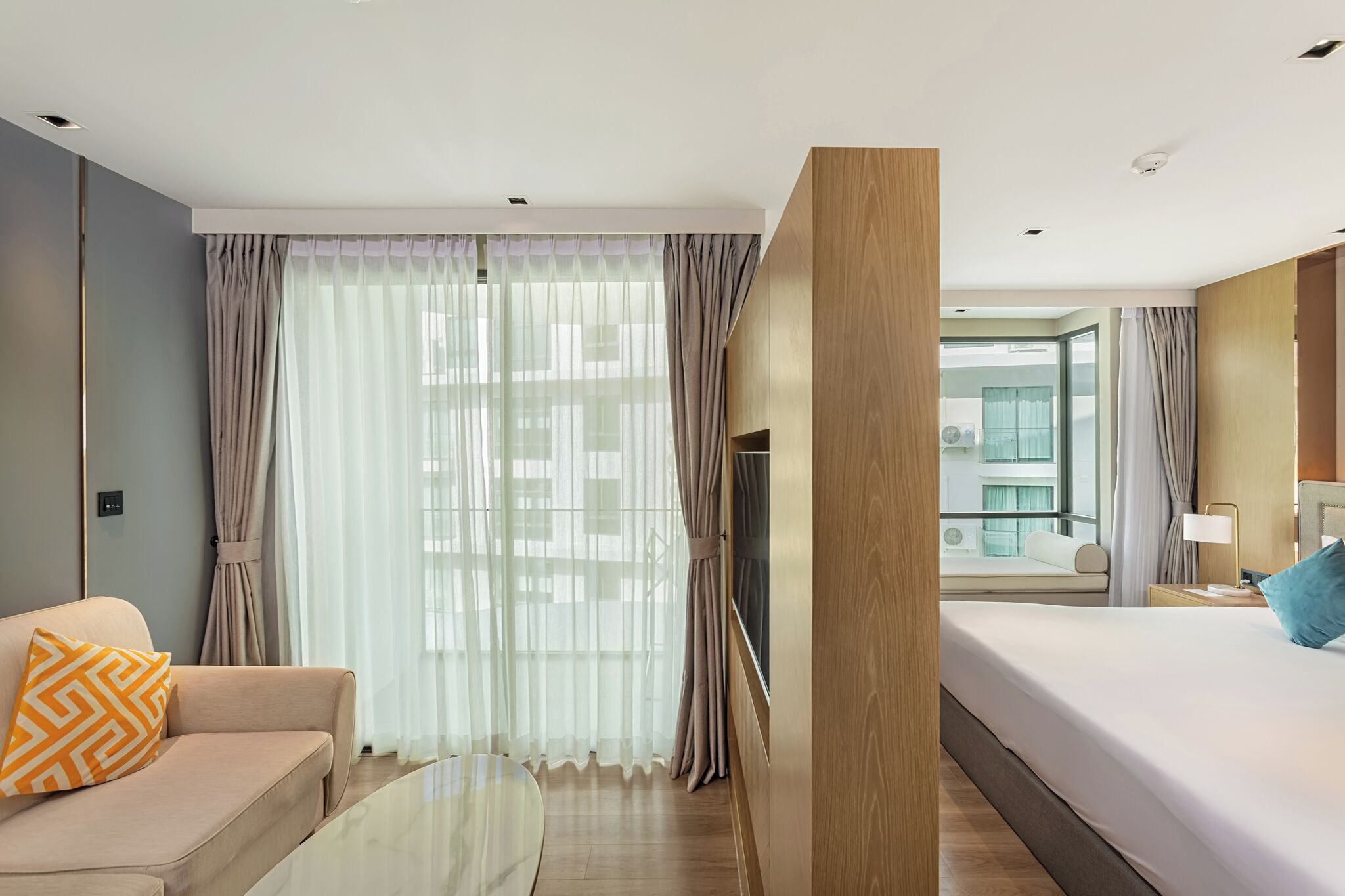 Une chambre d’hôtel moderne, dotée d’un grand lit, d’un canapé, d’une table basse en verre et offrant une vue sur la ville à travers de fines rideaux.