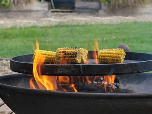 Ein Grill mit Maiskobs, Flammen und einem grasbewachsen Hintergrund.