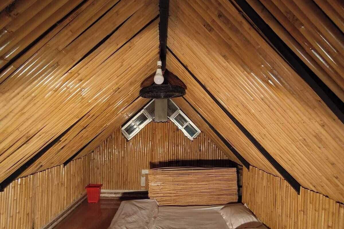 A small, cozy attic bedroom with a sloped wooden ceiling, a single bed with pillows, a red bucket, and a small table with flowers.