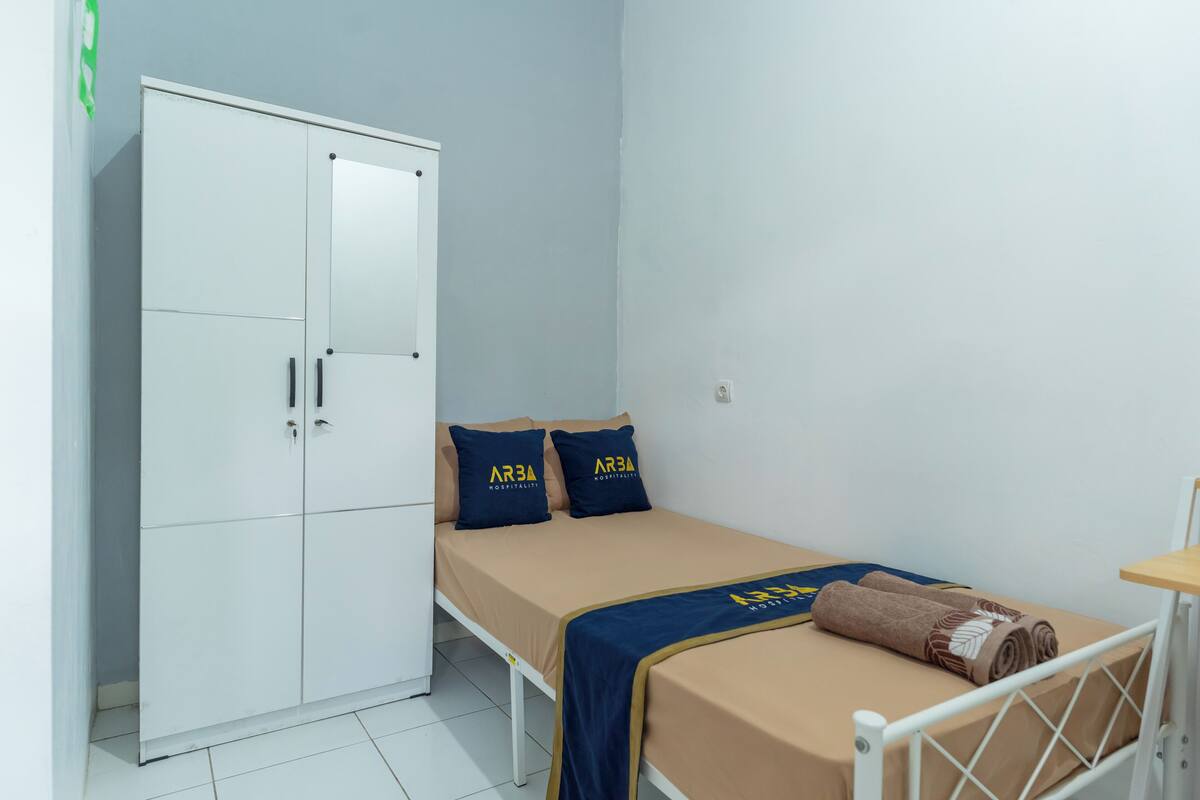 Een enkel bed met een beige matras en een blauwe deken, twee kussens met de tekst 'ARSA', een witte garderobe en een klein houten tafeltje.
