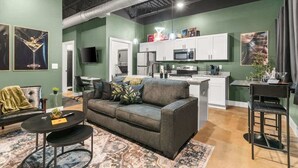 Un espacio habitable moderno con un sofá gris, una mesita redonda y una cocina con armarios blancos y un microondas.