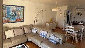A living room with a beige sectional sofa, a dining table with chairs, a painting on the wall, and a ceiling light.