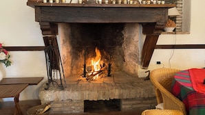 Una chimenea encendida, una repisa de madera con diversos objetos, un sillón de mimbre y una mesa con un florero.