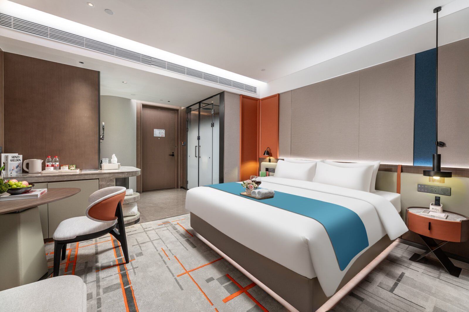 A modern hotel room with a large bed, a desk with a chair, a small table with a lamp, and a wardrobe.