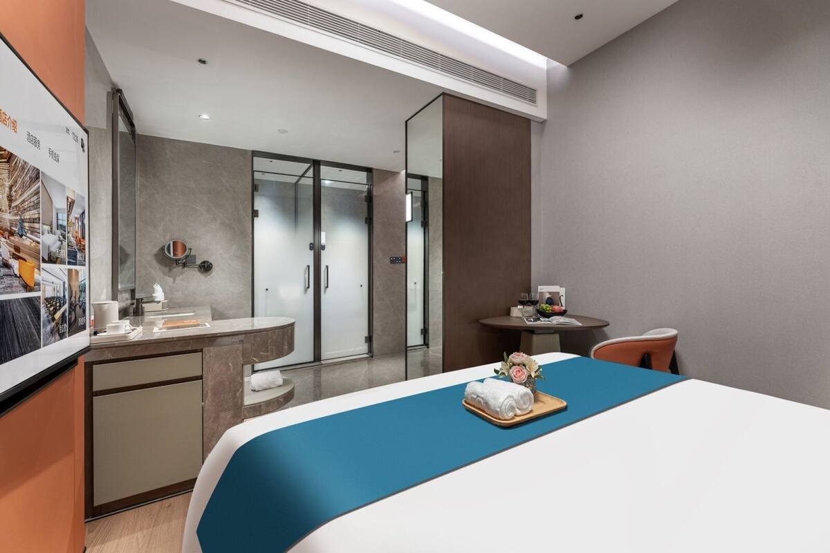 A hotel room with a bed, a desk, a chair, a small table with a bowl of fruit, and a mirror.