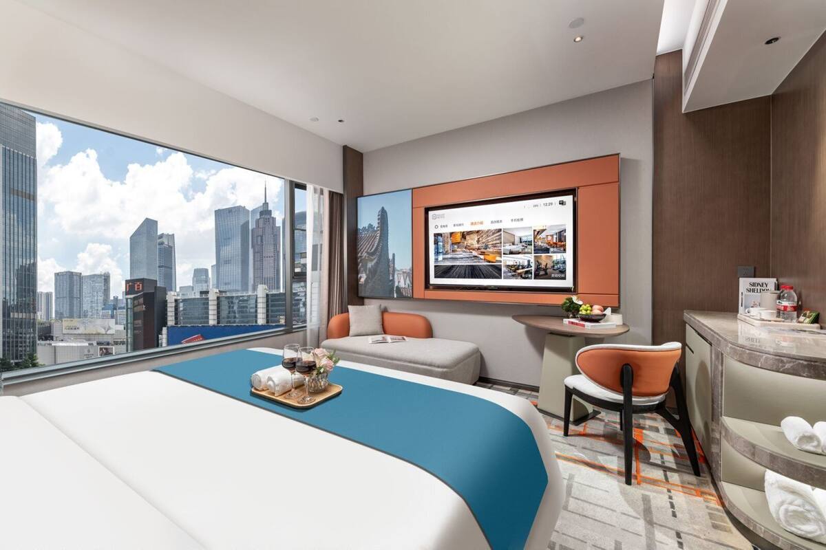 A modern hotel room with a large bed, a desk, a chair, a TV, and a city view.