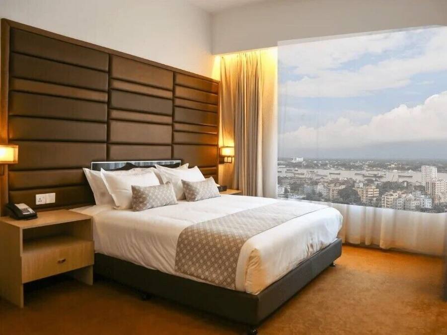 A hotel room with a large bed, a nightstand with a phone, and a view of a cityscape through a large window.