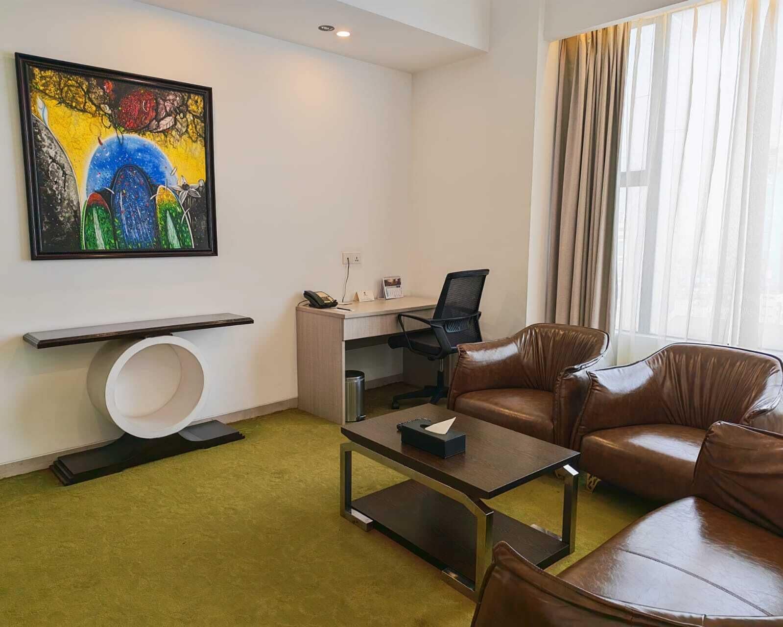 Una habitación de hotel moderna con un escritorio, sofá de cuero, una mesa de centro, un cuadro en la pared y un amplio ventanal con cortinas.
