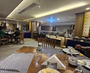 En restaurang med buffé, där gästerna kan välja mat från ett stort urval, med flera bord, ett centralt område där maten serveras och en dekorativ vägg med intrikata mönster.