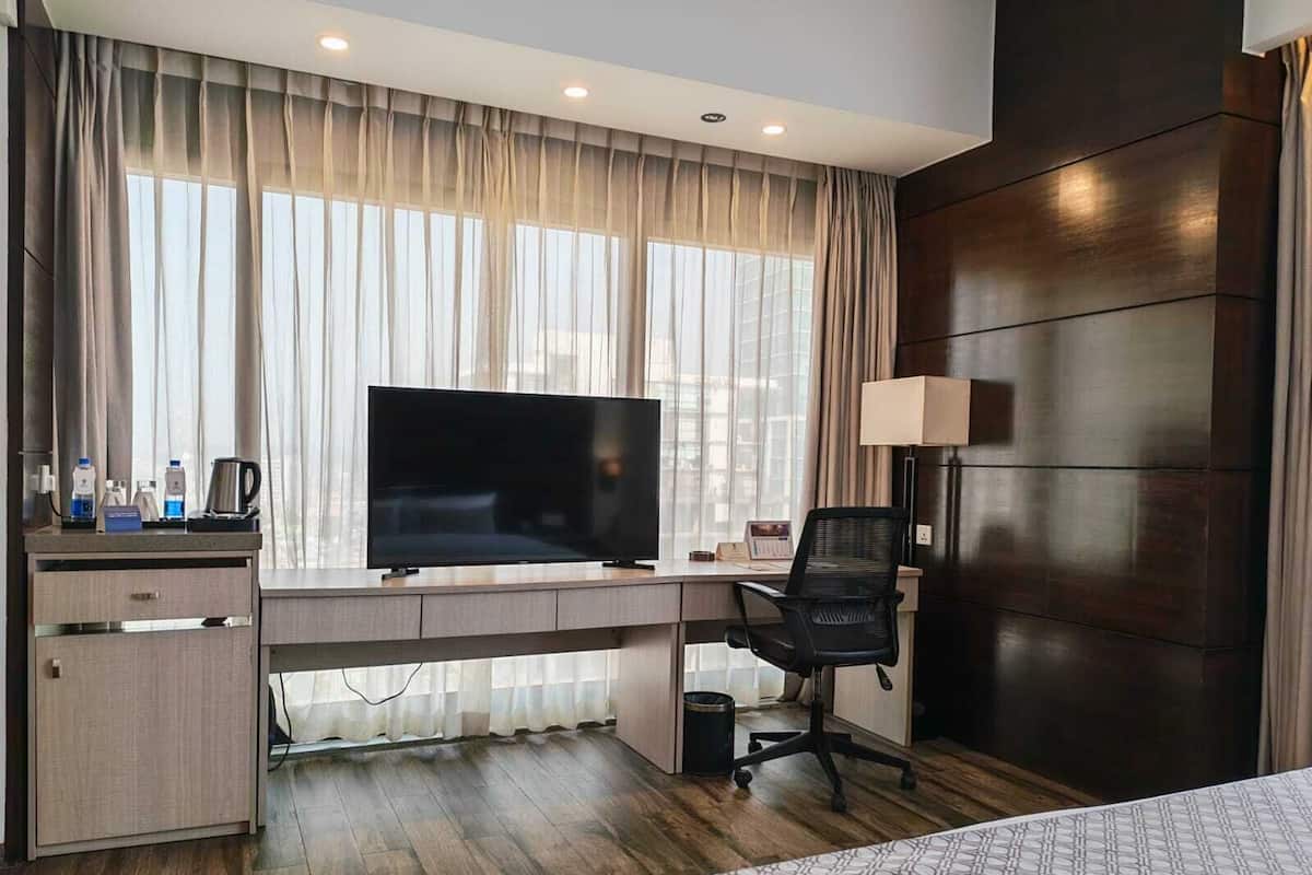 A hotel room with a large flat-screen TV, a desk with a chair, a bedside table, and a bed.