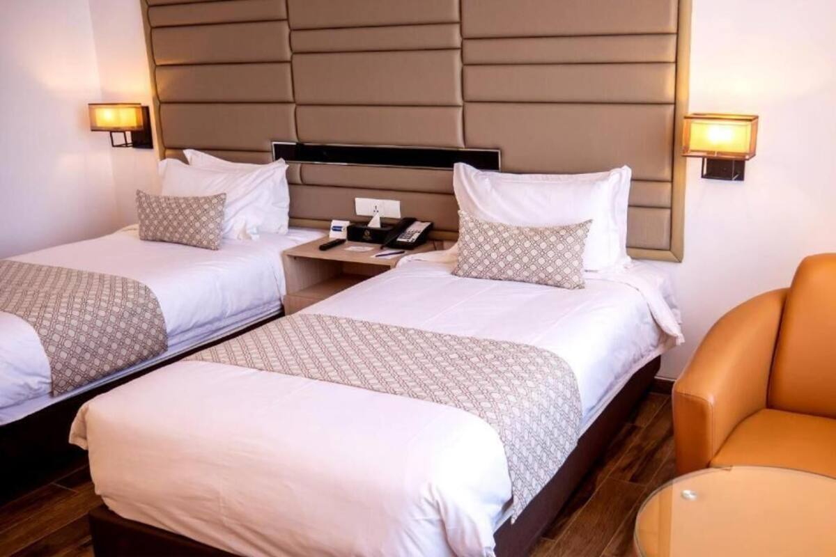A hotel room with two beds, a nightstand, a chair, and a wall-mounted lamp.