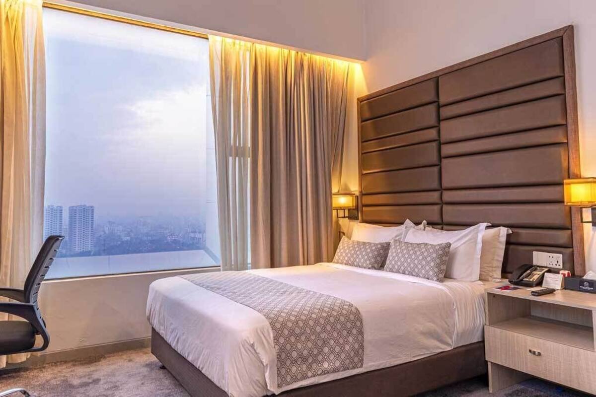 A hotel room with a large bed, a desk, a chair, a nightstand, and a view of the city.
