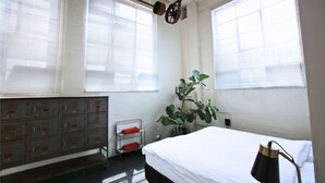 Una stanza con un letto, un armadietto metallico, una pianta in vaso e una finestra con persiane.