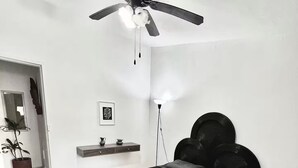 Un dormitorio con cama, ventilador de techo, lámpara de noche, una pequeña estantería y una planta en maceta.