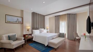 A hotel room with a bed, two chairs, a desk, a TV, and a painting on the wall.