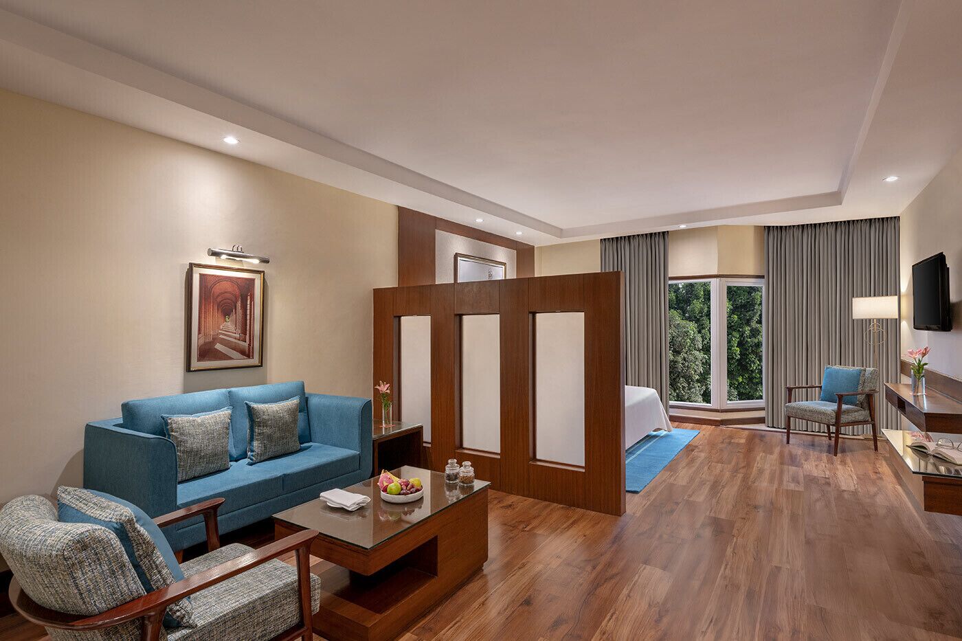 A modern hotel room with a blue sofa, a wooden coffee table, a small side table, a TV, and a large window with curtains.