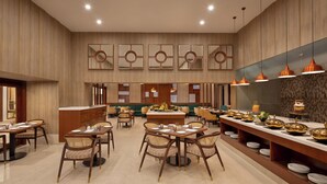 A spacious hotel dining area with multiple tables and chairs, a buffet counter, and decorative wall mirrors.