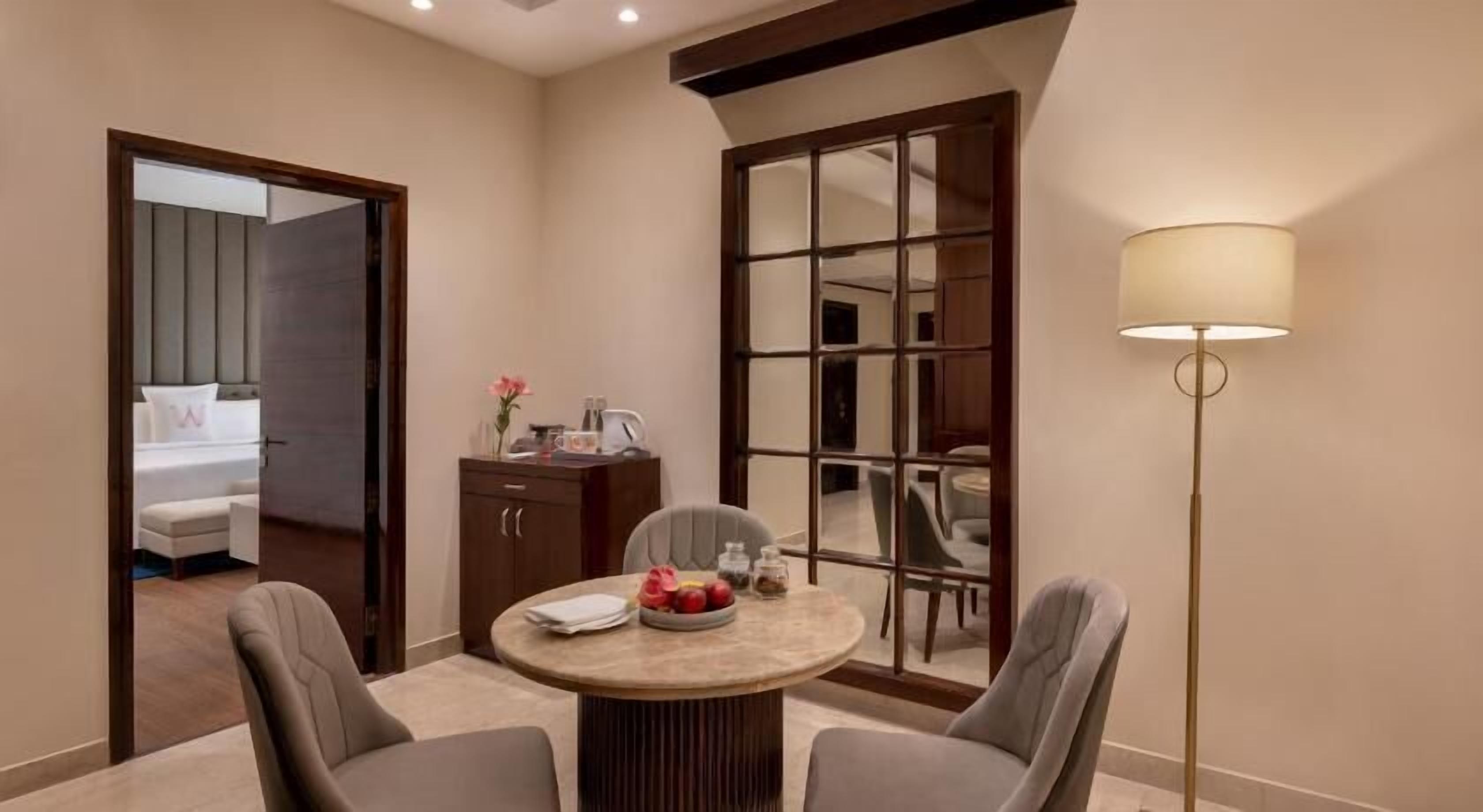 A hotel room with a round dining table, two chairs, a cabinet, a floor lamp, and a large mirror.