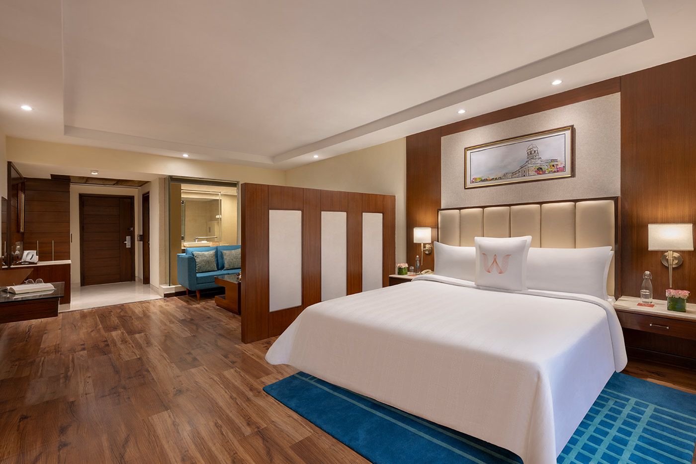 A hotel room with a large bed, a wooden floor, a blue rug, a nightstand with a lamp, and a wall-mounted painting.
