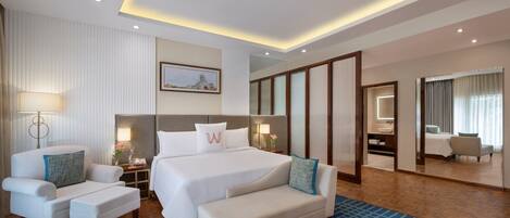 A modern hotel room with a large bed, two armchairs, a sofa, a bedside table, a lamp, a mirror, and a view of the bathroom.