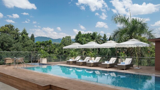 A swimming pool with lounge chairs and umbrellas, surrounded by greenery and a view of mountains.