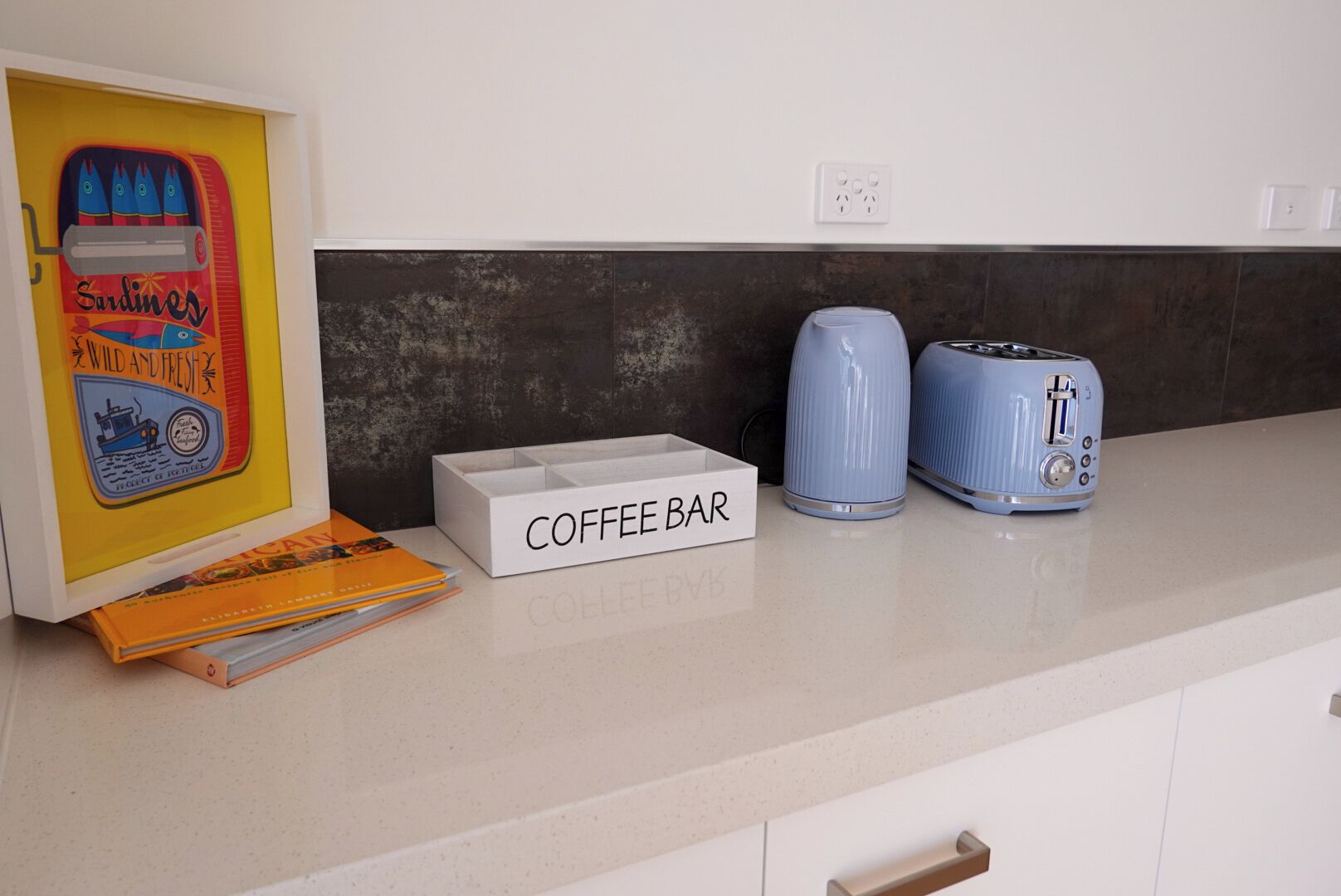 Una encimera de cocina con una estación para café, que incluye una caja blanca con la etiqueta 'COFFEE BAR', una tetera azul y un tostador azul.