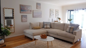 A modern living room with a beige sectional sofa, a small white coffee table, and a large mirror. There are several framed artworks on the walls and a potted plant on the floor. - Casa Mar - Beachside Bliss.
Sun, sand and family fun All in one place. (Carrum)
