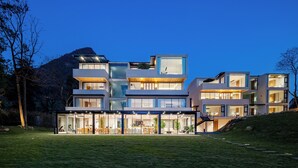 Une maison moderne à plusieurs étages, dotée de grandes fenêtres en verre et d’un toit plat, entourée d’une pelouse et d’arbres.