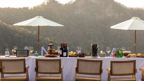 Une table dressée pour un repas, avec des bouteilles de vin, des verres et des fruits, donnant sur une colline boisée.