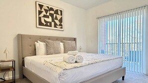 Une chambre à coucher avec un grand lit, une tête de lit, une table de chevet, une lampe et une fenêtre avec des stores.