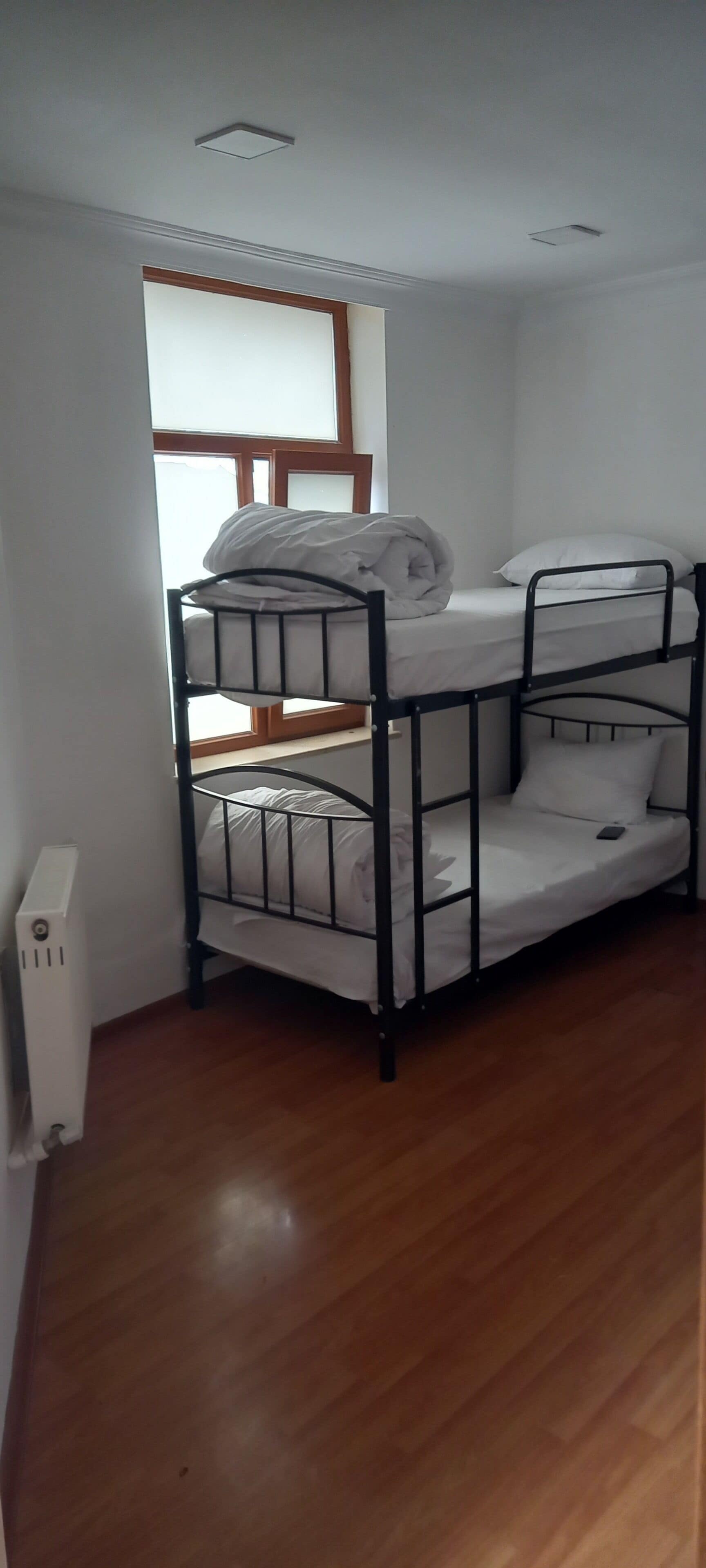 A bunk bed room with white bedding, a radiator, and a window with blinds.
