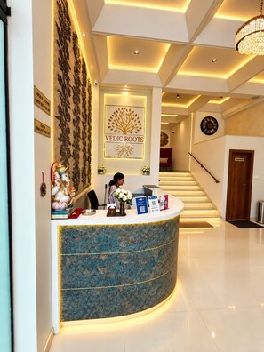 A reception desk with a logo 'Vedic Roots' and a receptionist. There is a staircase in the background. - Hotel Vedic Roots (Varanasi)