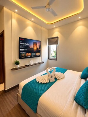 A hotel room with a bed, a television mounted on the wall, a ceiling fan, and a window with a view. - Hotel Vedic Roots (Varanasi)