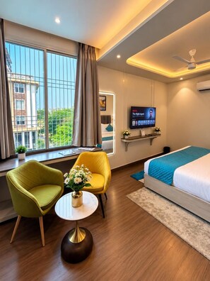 A hotel room with a bed, a television, a window with curtains, a small table with a vase, and two chairs. - Hotel Vedic Roots (Varanasi)