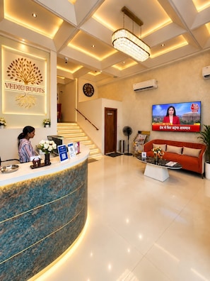 A reception area with a reception desk, a chandelier, a television displaying news, and a staircase in the background. - Hotel Vedic Roots (Varanasi)