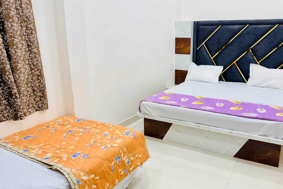 Ein Schlafzimmer mit einem Bett, das ein dunkelbraunes Kopfteil mit geometrischen Mustern, eine weiße Matratze und eine violette Decke mit floralen Motiven aufweist. Es gibt ein weiteres Bett mit einer orangefarbenen, blumengemusterten Tagesdecke und einem braunen Vorhang.