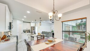A modern kitchen with white cabinets, a wooden dining table, and a large window with blinds. - Heated Pool & Outdoor Kitchen: Cape Coral Home (Cape Coral)