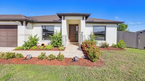 A two-story house with a front entrance, a garage, and a well-maintained lawn with small shrubs and a flower bed. - Heated Pool & Outdoor Kitchen: Cape Coral Home (Cape Coral)