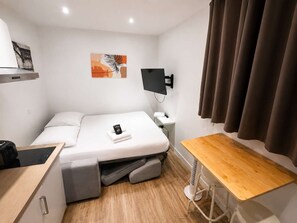 Een kleine hotelkamer met een bed, een bureau, een bijzettafeltje, een tv aan de muur en een stopcontact.