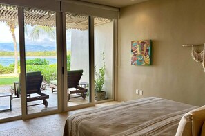 A bedroom with a bed, a sliding glass door leading to a balcony with a chair and a plant, and a colorful artwork on the wall. - Dream Villa in Majahua (La Majahua)