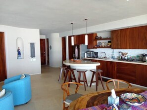 A modern kitchen with wooden cabinets, a central island with stools, and a dining area with a round table and blue chairs. - Dream Villa in Majahua (La Majahua)