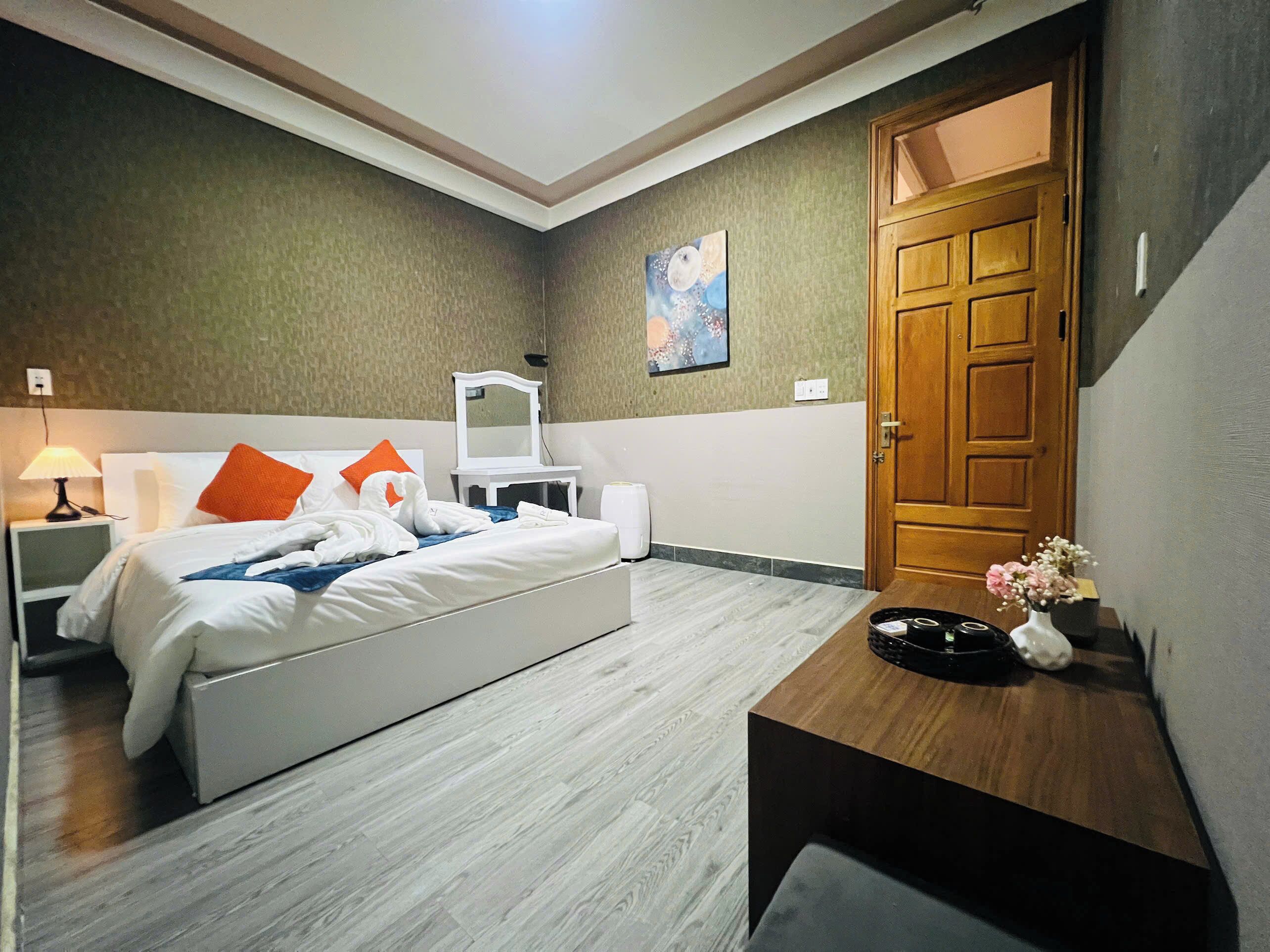 A hotel room with a bed, a desk, a mirror, a painting, and a wooden door.