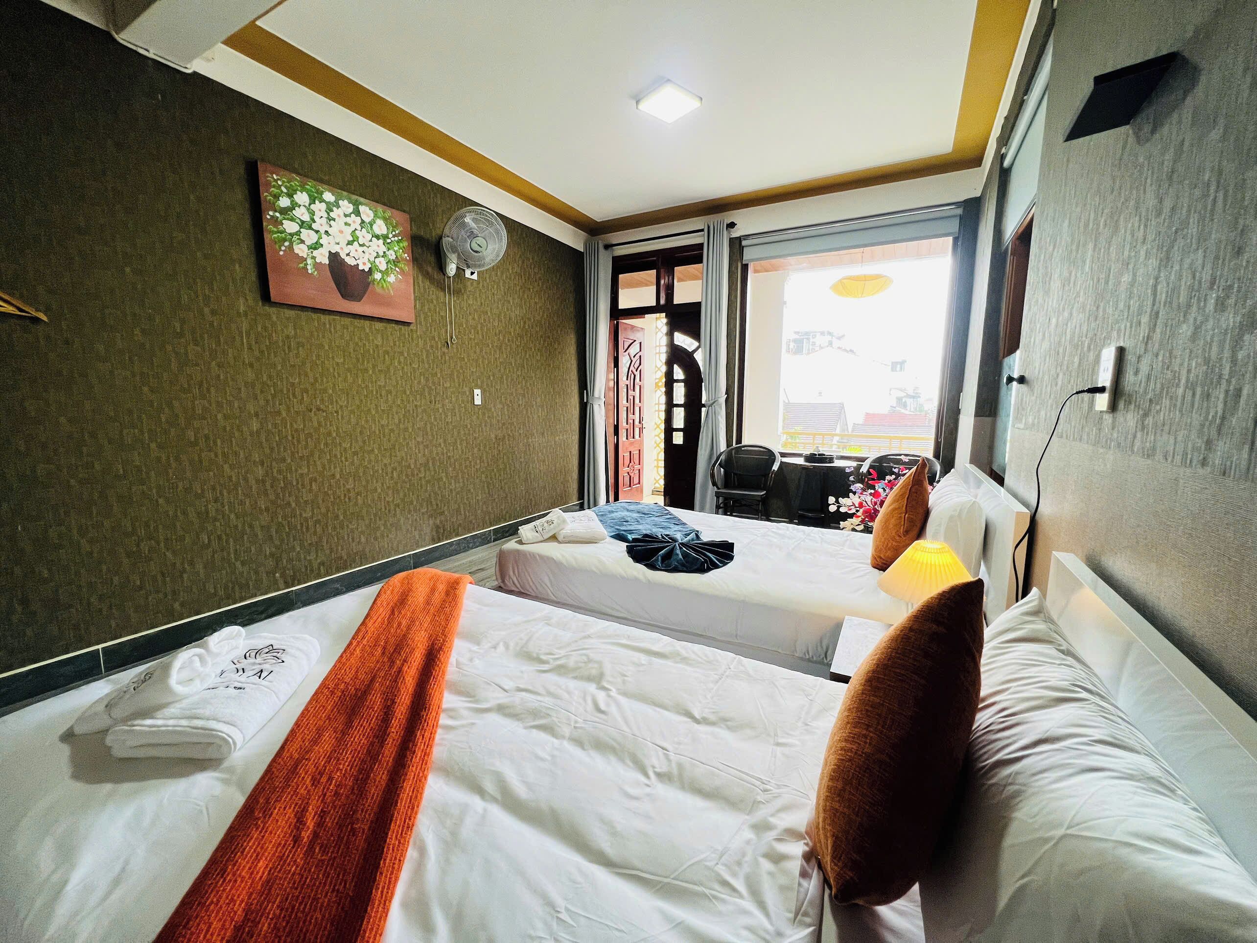 A hotel room with two beds, a painting, a fan, and a view of the outside.