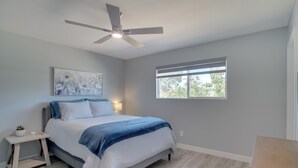 Un dormitorio con cama, ventilador de techo, ventana y mesita de noche.