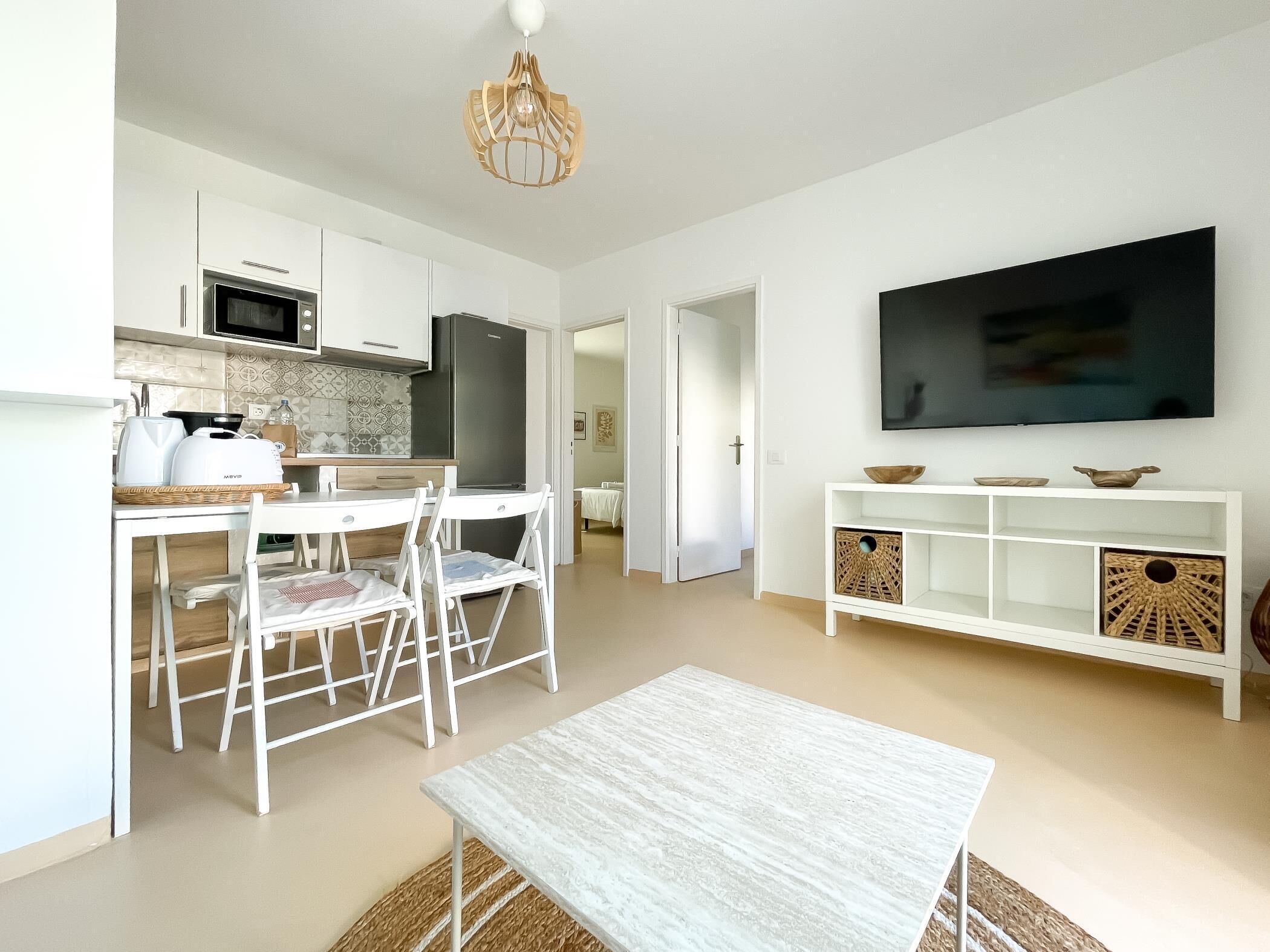 Eine moderne Küche mit weißen Schränken, einer Mikrowelle und einem Kühlschrank. Ein Essbereich mit weißem Tisch und Stühlen ist ebenfalls vorhanden. Ein Flachbildfernseher ist an der Wand oberhalb eines weißen Konsolentisches mit Dekorationen angebracht.