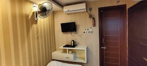 Ein Hotelzimmer mit einem Bett, einem an der Wand montierten Fernseher, einem kleinen Tisch mit einem Wasserkocher, einem Ventilator und einer Tür.