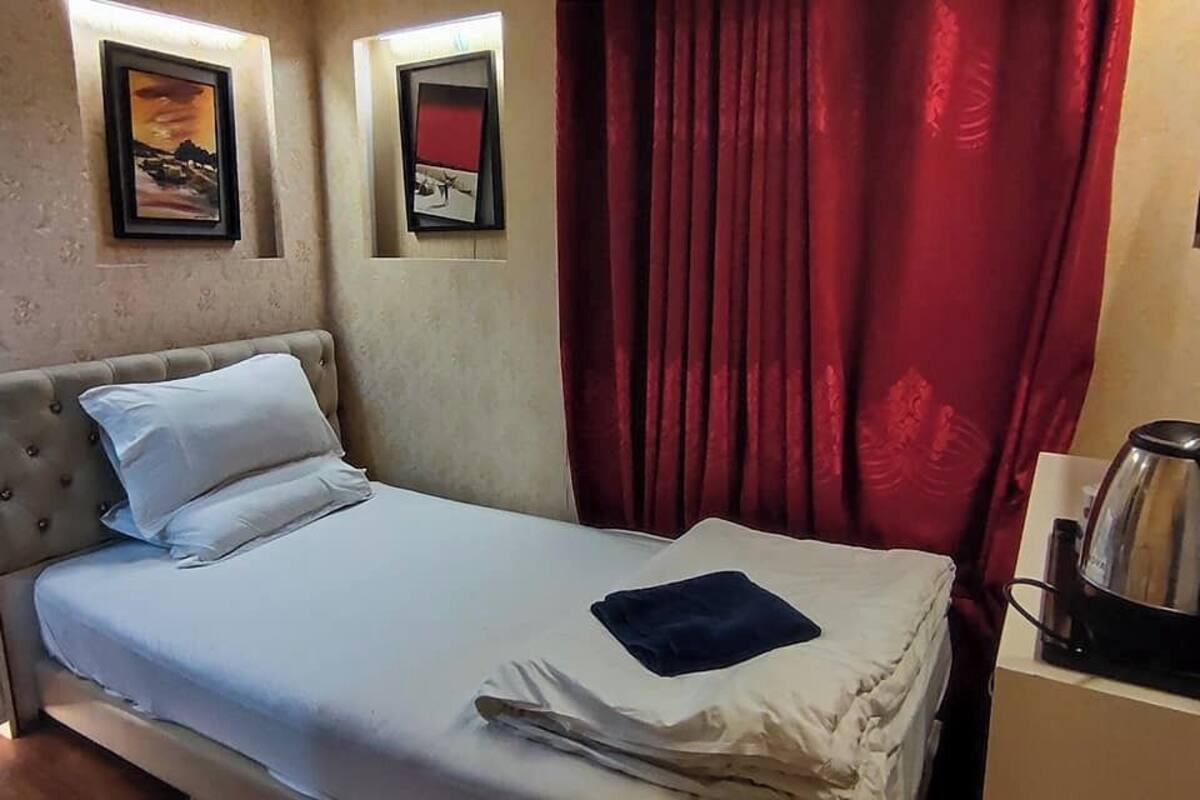 Habitación de hotel con cama, mesita de noche, lámpara, cafetera y dos cuadros en la pared.