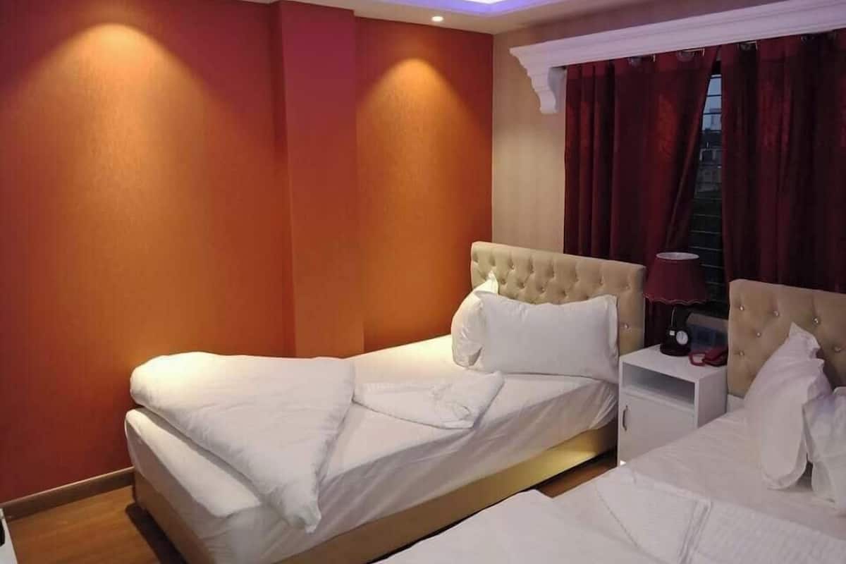 Habitación de hotel con dos camas, una mesita de noche, una lámpara y una ventana con cortinas.