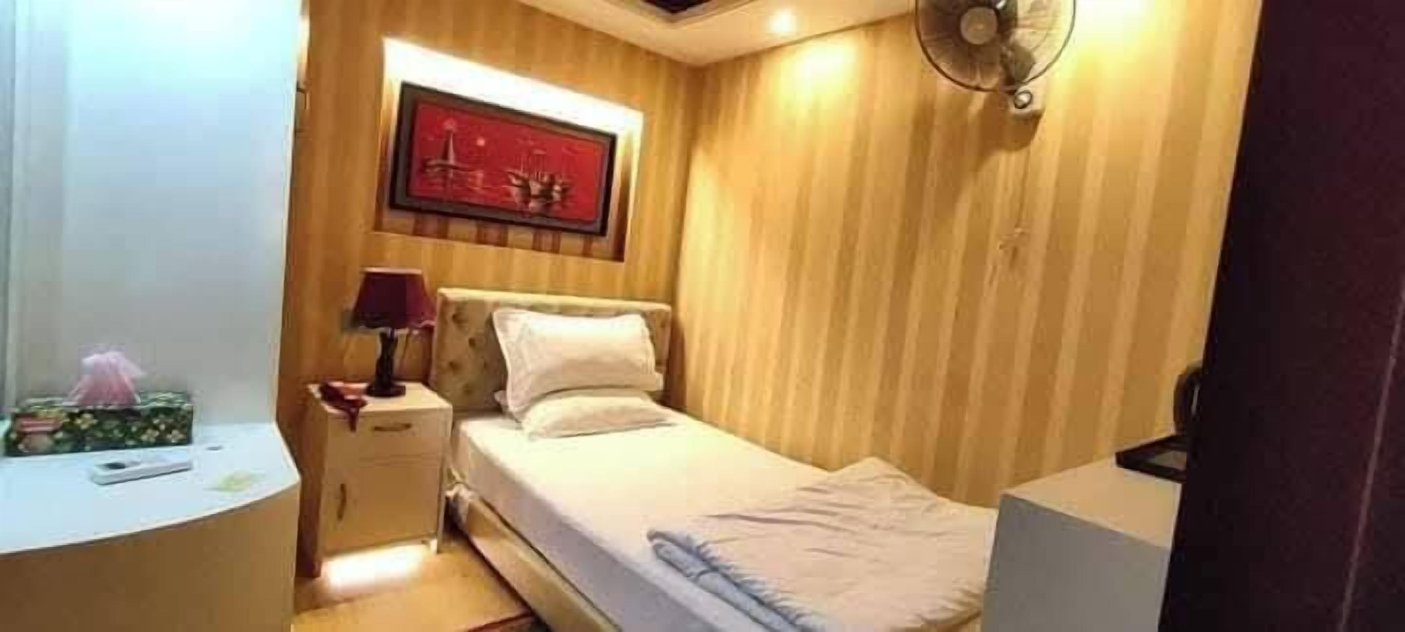 A small, neatly arranged bedroom with a bed, bedside table, lamp, and a framed picture on the wall.